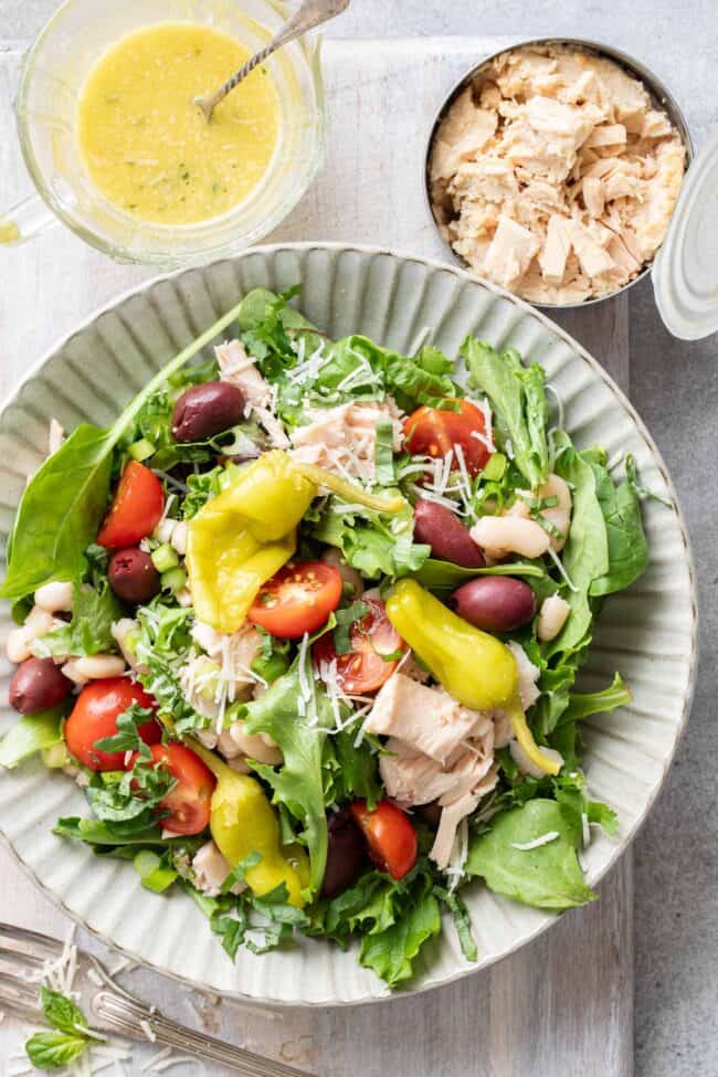 A grey plate filled with white bean tuna salad. An opened can of tuna and glass jar filled with salad dressing sits next to the plate.