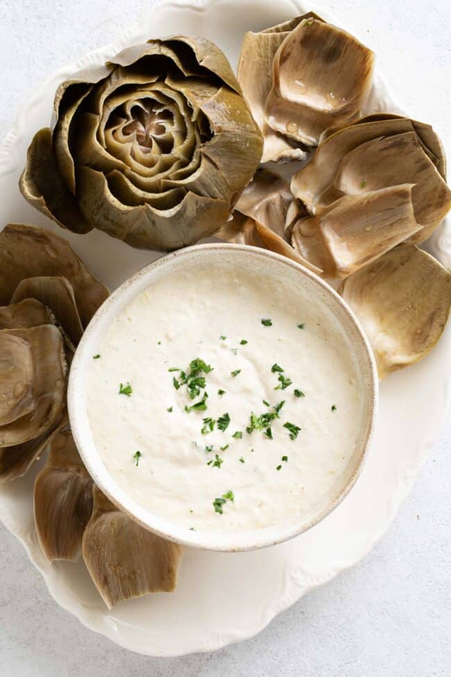 A cooked artichoke with scattered artichoke leaves and a bowl filled with a creamy dip sit on a white platter.