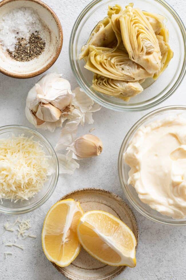 Glass measuring bowls filled with mayonnaise, marinated artichoke hearts, grated parmesan cheese, lemon wedges and garlic cloves.