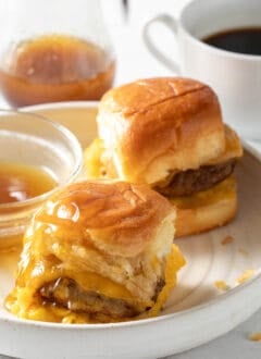 A white plate with two breakfast sliders on it next to a small bowl of maple syrup.