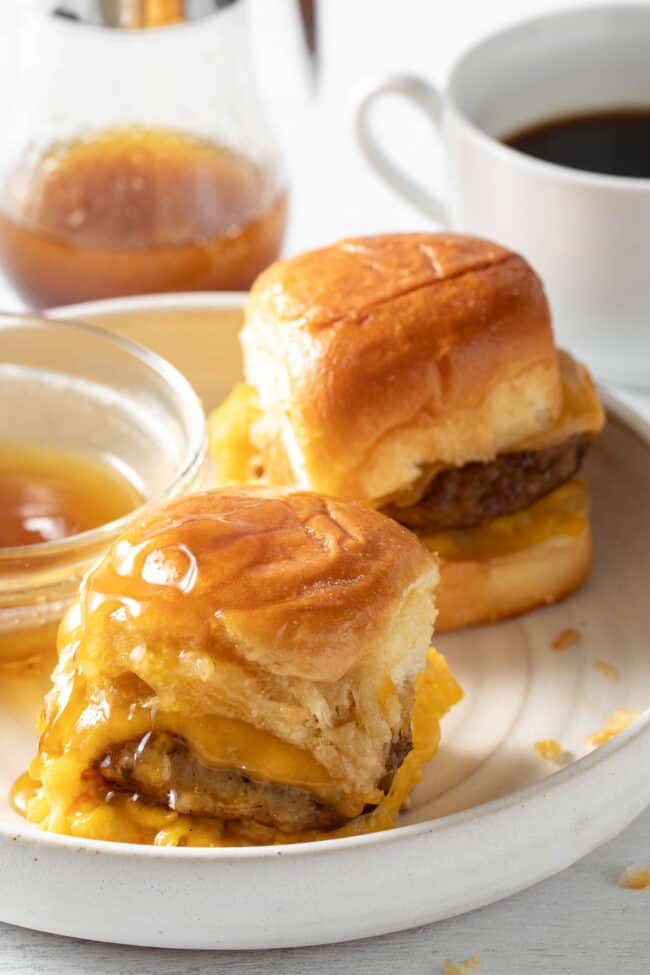 A white plate with two breakfast sliders on it next to a small bowl of maple syrup.