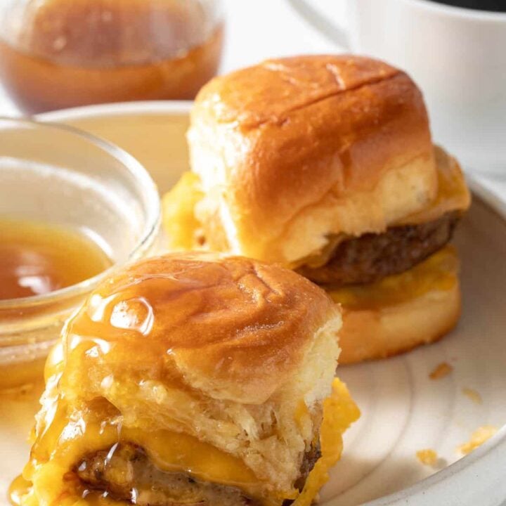 A white plate with two breakfast sliders on it next to a small bowl of maple syrup.