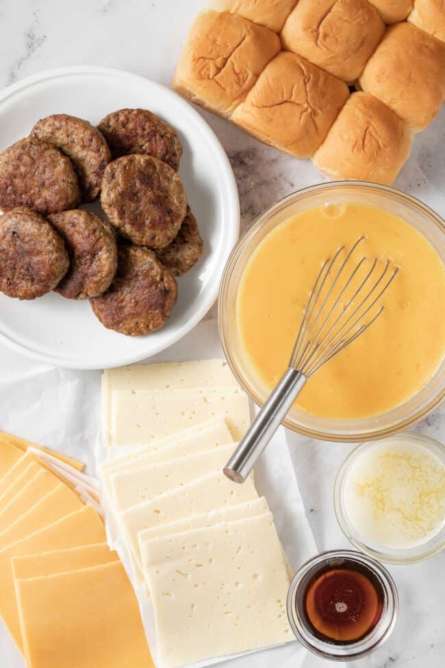 A plate with cooked sausage sits next to a bowl of raw eggs with a whisk, slices of cheese and Hawaiian rolls.