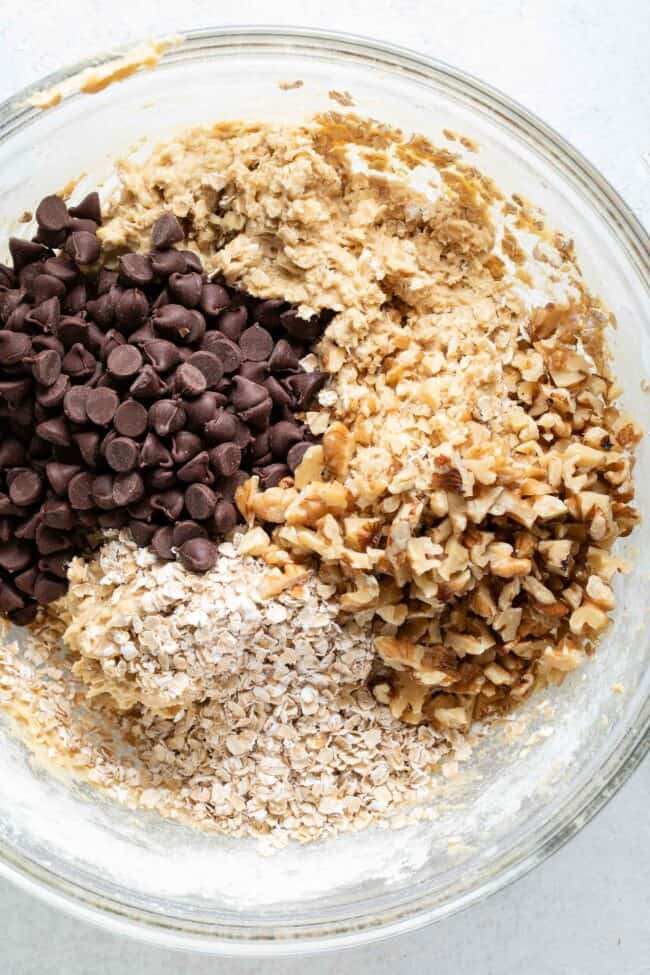 A clear glass mixing bowl filled with batter, chocolate chips, walnuts and oats. 