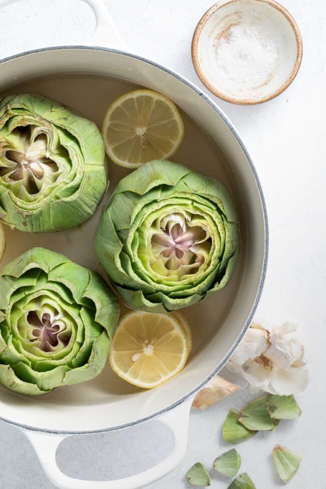 Three large artichokes in a white Dutch oven filled halfway with water.