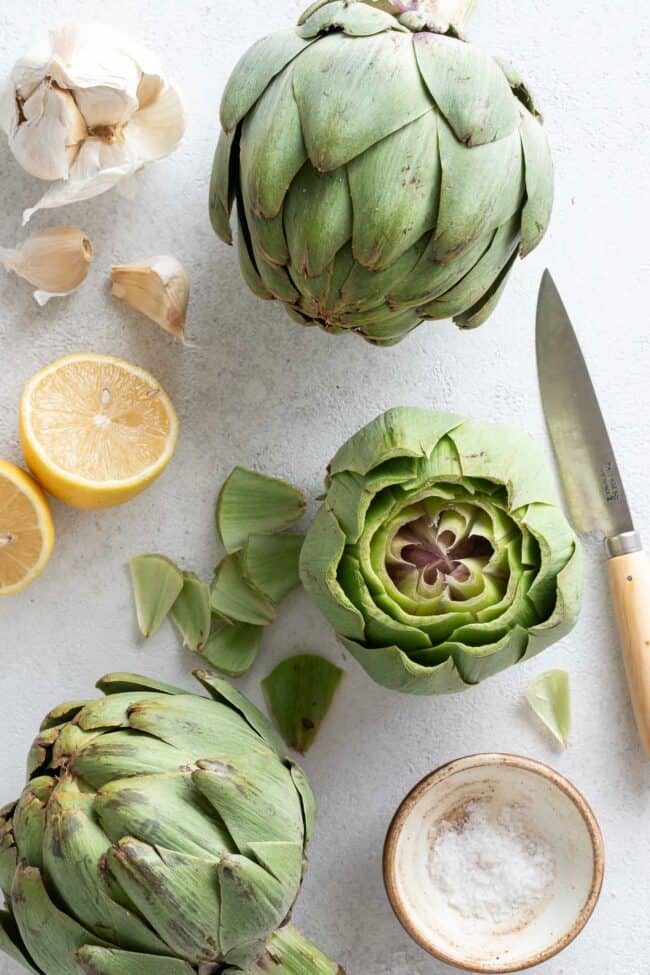 Green vegetables next to a lemon cut in half, garlic cloves and a knife.