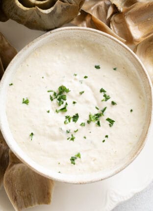 A white bowl filled with artichoke dipping sauce. Artichoke leaves are scattered around the bowl.