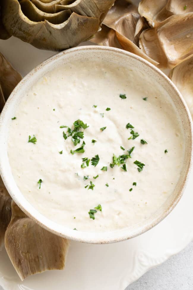 A white bowl filled with artichoke dipping sauce. Artichoke leaves are scattered around the bowl.