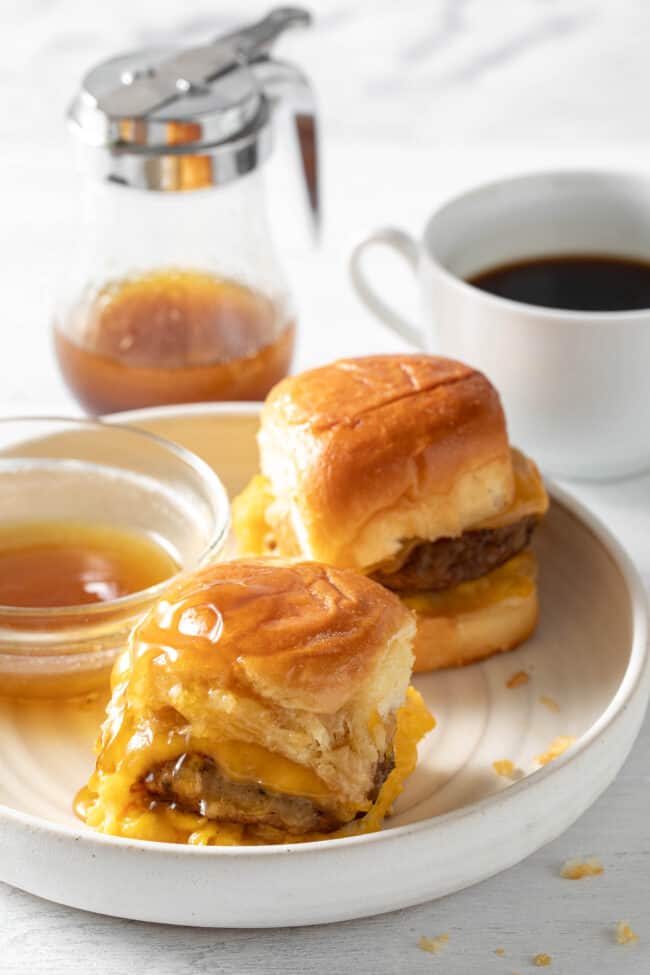 A white plate with two breakfast sliders on it next to a small bowl of maple syrup.