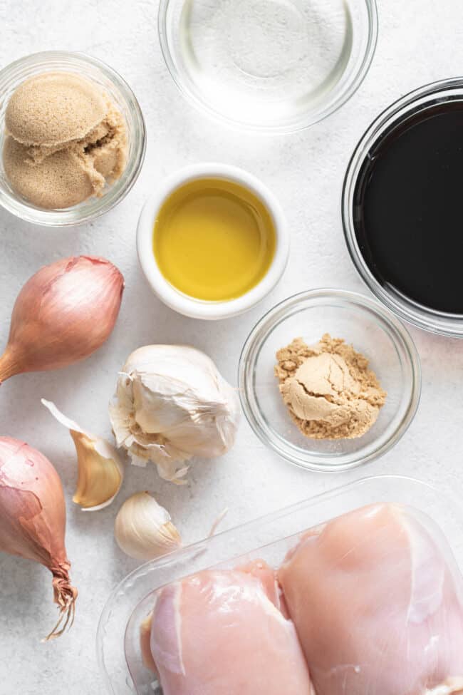 Clear glass measuring bowls with vinegar, soy sauce, olive oil, brown sugar and powdered ginger. Two raw chicken breasts sit next to the ingredients.