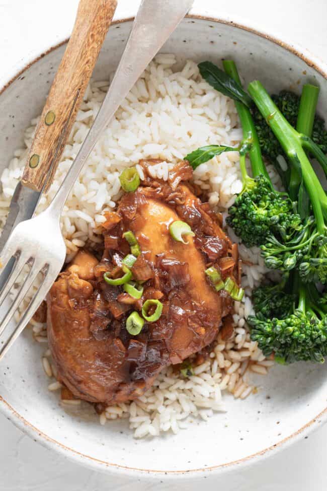 A white plate with Filipino Adobo chicken on a bed of white rice. Steamed broccoli sits on the plate next to the chicken.