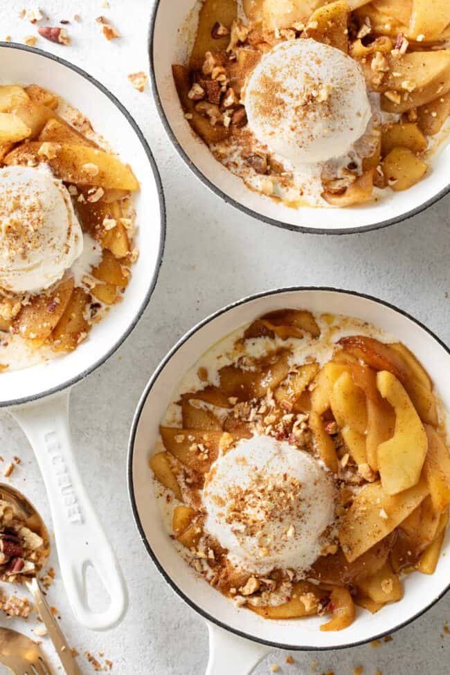 Three small white skillets filled with fried apples and scoops of vanilla ice cream.