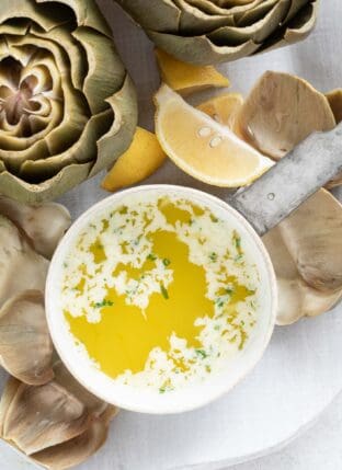 A small white saucepan filled with garlic butter sauce. Artichoke leaves are scattered around the saucepan.