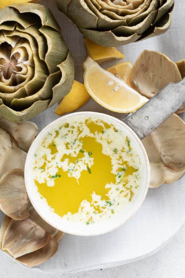 A small white saucepan filled with garlic butter sauce. Artichoke leaves are scattered around the saucepan.