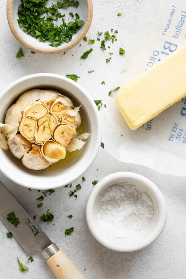 A stick of butter sits next to three small bowls filled with a head of roasted garlic, salt and chopped parsley.