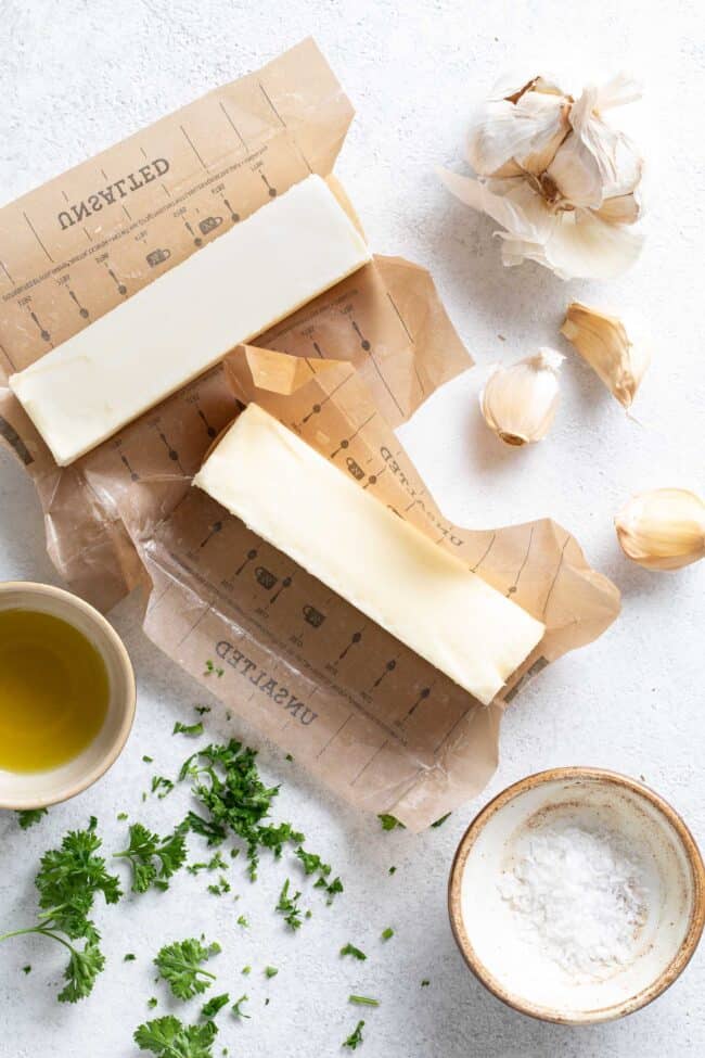 Two sticks of butter next to garlic cloves, chopped parsley and a small bowl with olive oil in it.