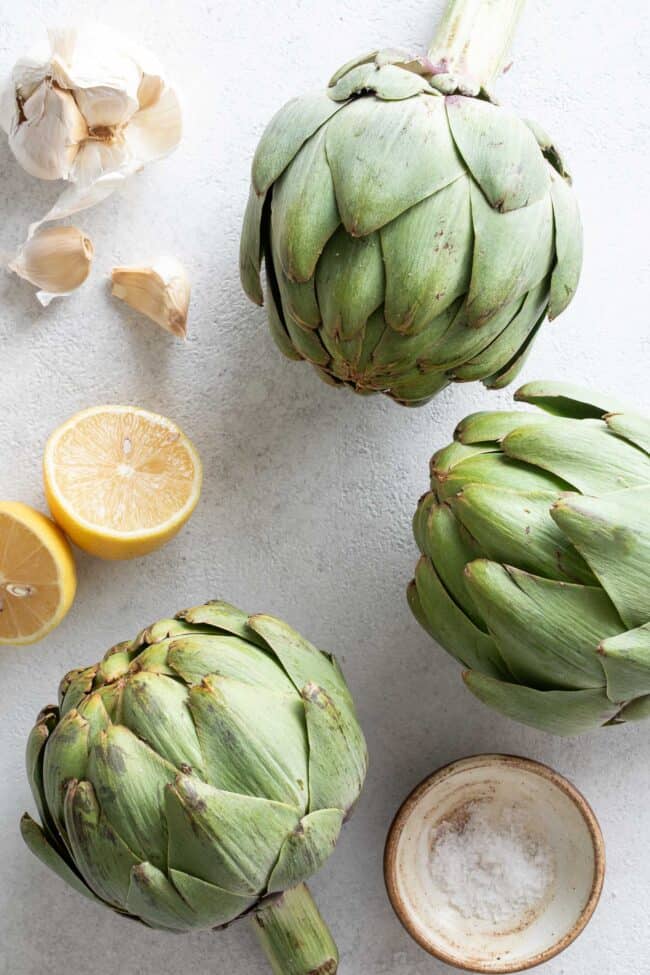 Three artichokes next to a lemon cut in half and garlic cloves for steamed artichokes recipe.