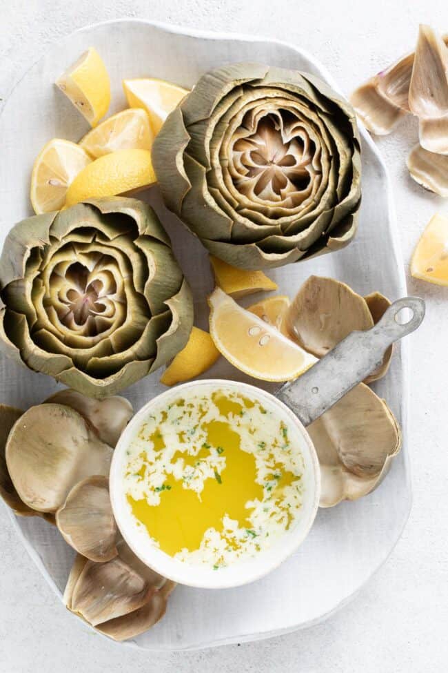 A platter with two cooked artichokes. A small saucepan filled with a dip sits next to the artichokes.