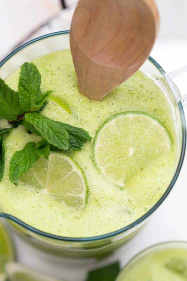 A clear glass pitcher filled with a green drink. Garnished with lime slices and fresh mint.