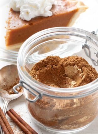 A mason jar filled with homemade pumpkin pie spice. A piece of pumpkin pie sits behind the jar.