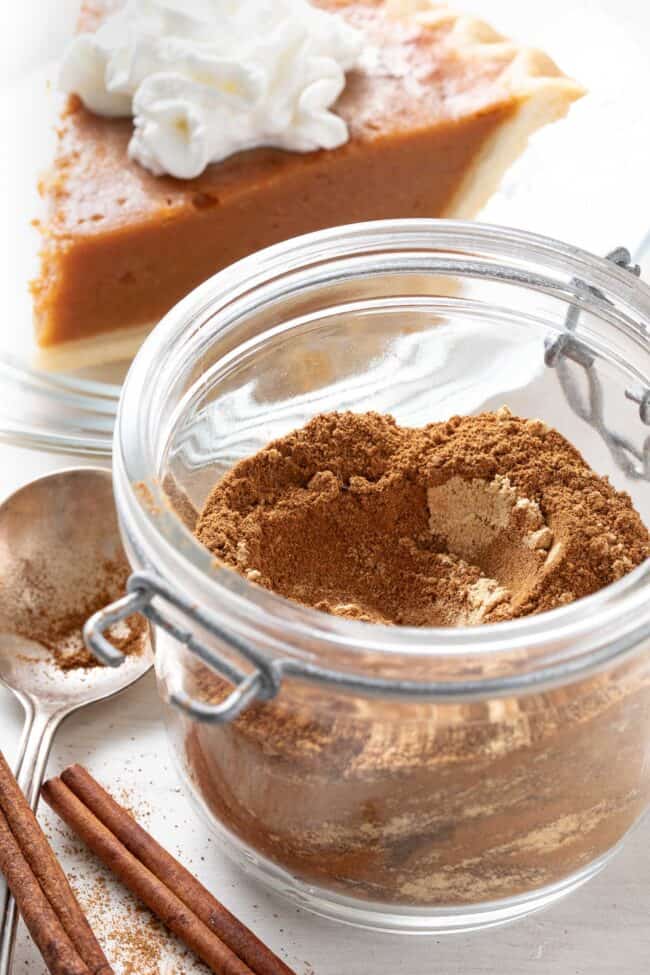 A mason jar filled with homemade pumpkin pie spice. A piece of pumpkin pie sits behind the jar.