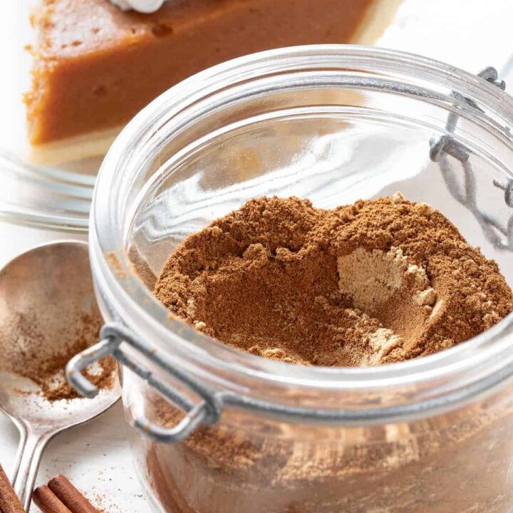 A mason jar filled with homemade pumpkin pie spice. A piece of pumpkin pie sits behind the jar.