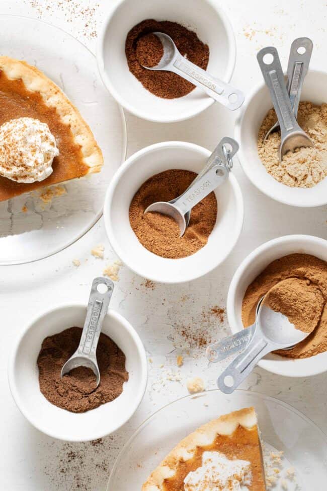 Five small bowls filled with pumpkin pie spices. Two pieces of pumpkin pie with whipped cream sit next to the bowls.