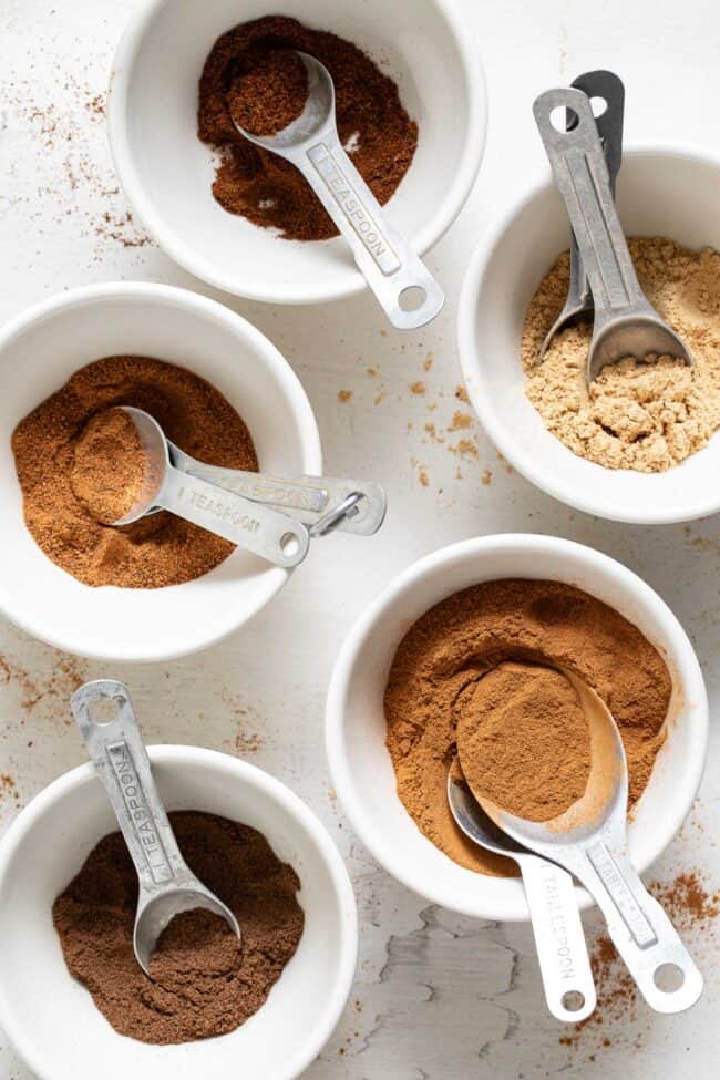 Five small white bowls filled with cinnamon, nutmeg, ginger, cloves and allspice.