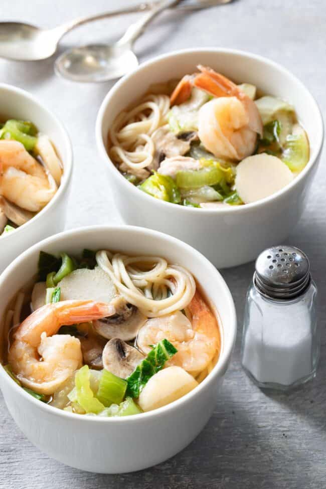 Two white bowls filled with fresh vegetables and meats in broth.