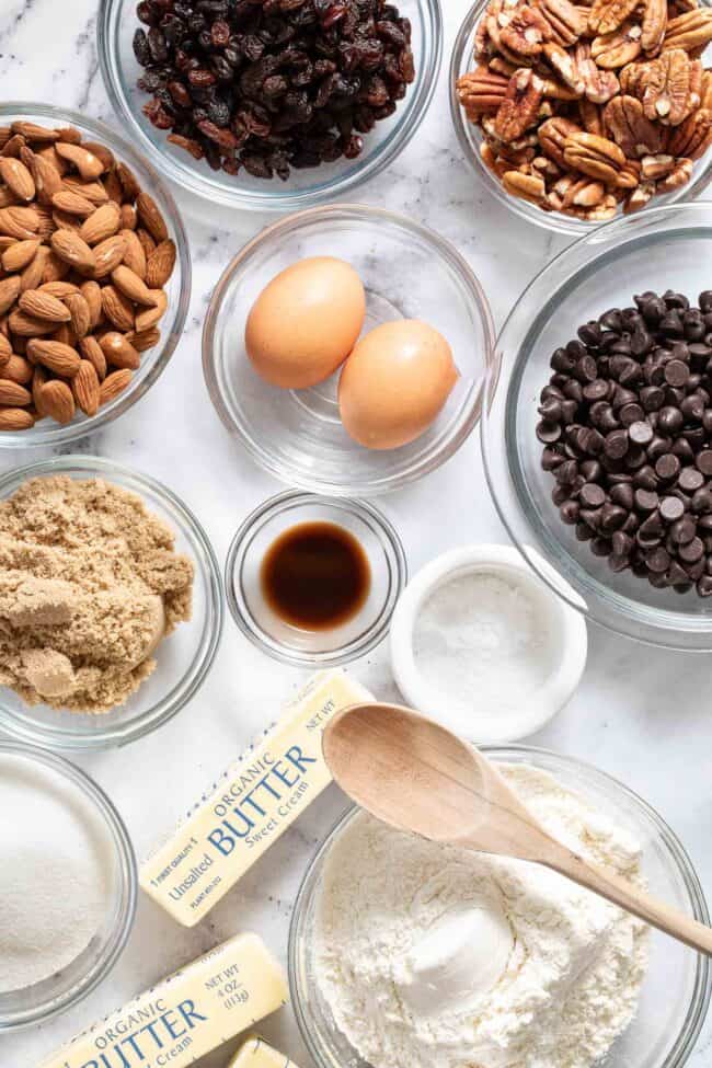 Clear glass mixing bowls filled with nuts, chocolate chips, brown sugar, granulated sugar and flour.