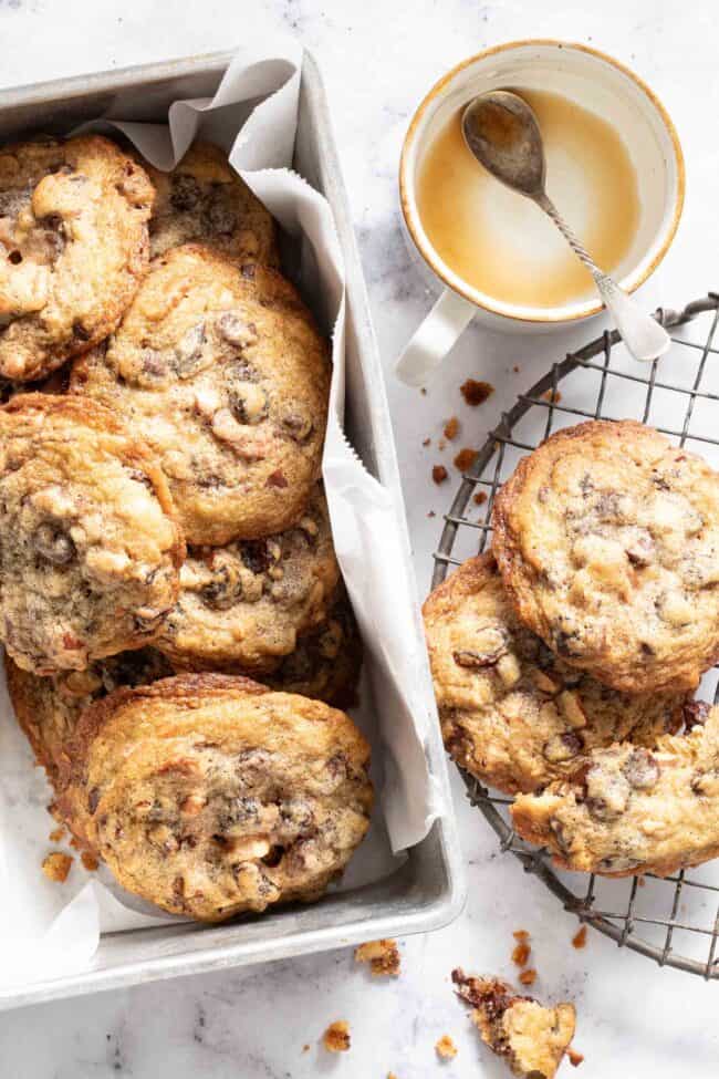 An aluminum bread tin filled with cowboy cookies. An empty coffee cup sits next to the tin.