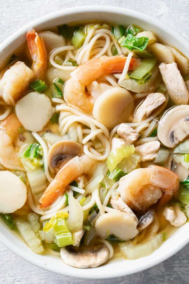 A large white bowl filled with noodles, cooked shrimp and chicken and vegetables in broth.