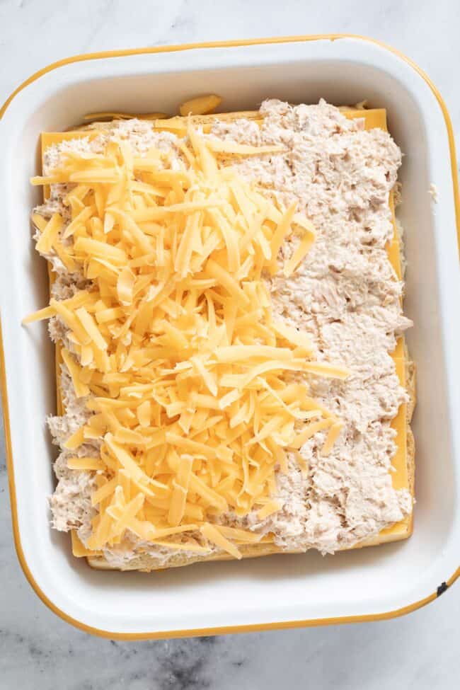 A white baking dish with tuna salad and grated cheddar cheese on top of Hawaiian rolls for tuna melt sliders.
