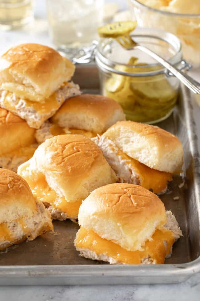 A cookie sheet filled with tuna melt sliders. A small jar of pickle slices sits on the tray with the sliders.