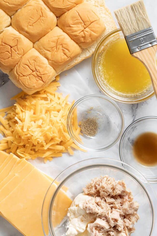 A package of Hawaiian rolls sits next to grated cheese, cheddar cheese slices and a clear glass mixing bowl of canned tuna with mayonnaise.