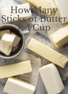 Several sticks of butter and wrappers sit next to an aluminum measuring cup filled with butter.