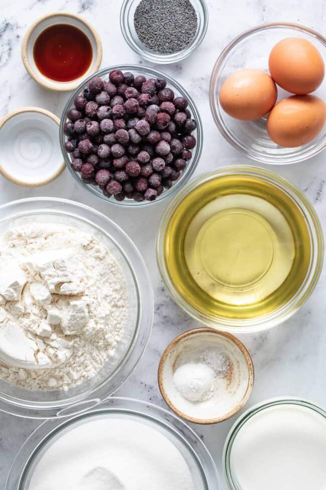 Clear glass mixing bowls filled with flour, sugar, salt, blueberries, eggs and oil.