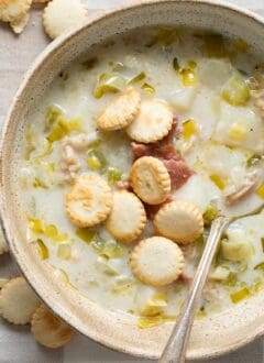 A tan colored bowl filled with New England Clam Chowder topped with pieces of bacon and oyster crackers.