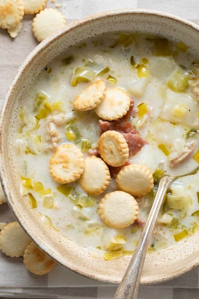 A tan colored bowl filled with New England Clam Chowder topped with pieces of bacon and oyster crackers.