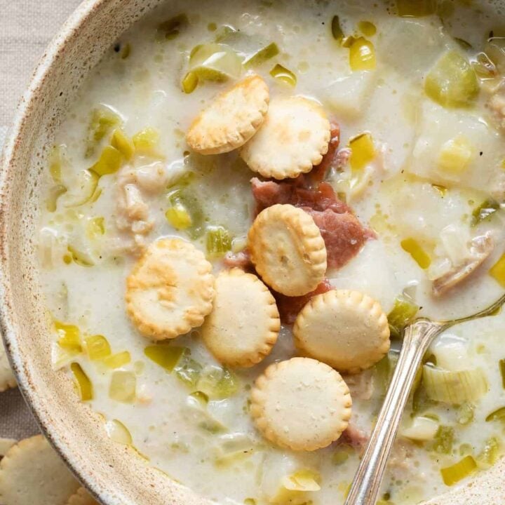 A tan colored bowl filled with New England Clam Chowder topped with pieces of bacon and oyster crackers.