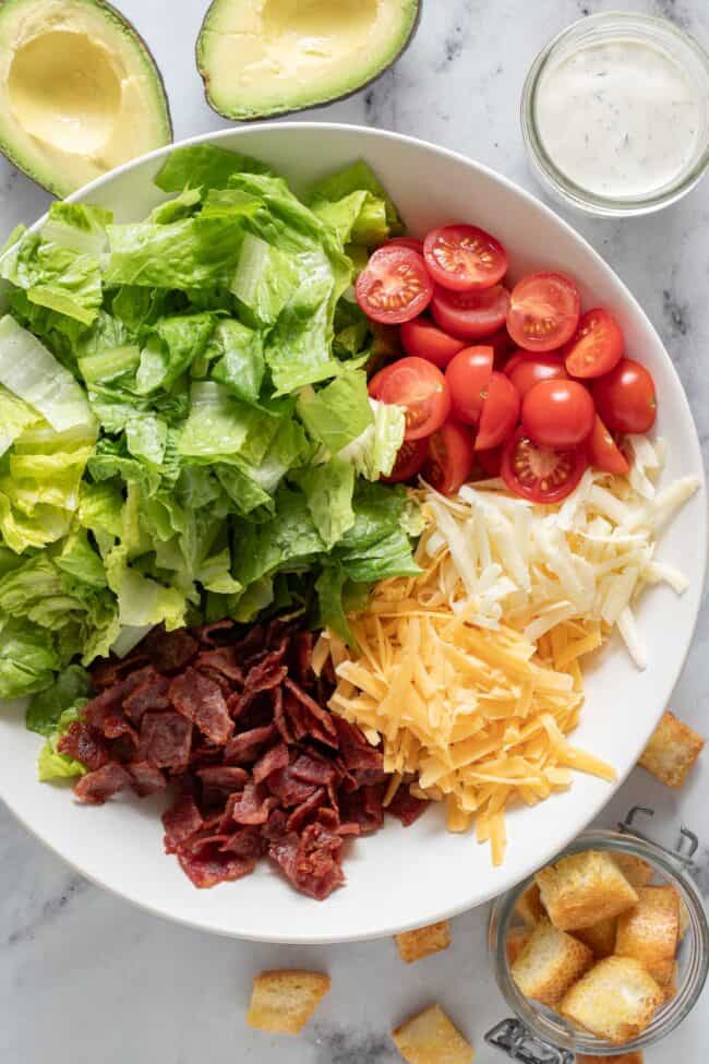 A white bowl filled with chopped lettuce, tomatoes, cooked bacon and grated cheese.