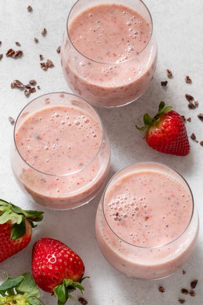 An overhead photo of three clear glasses filled with Strawberry Cacao Smoothie. Fresh strawberries and cacao nibs are scattered around the glasses.