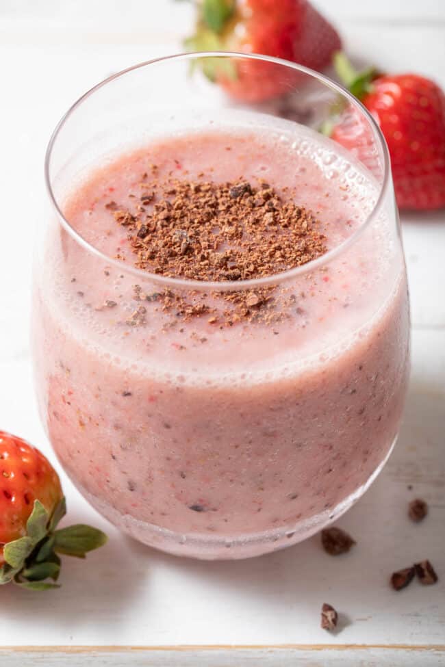 A clear glass filled with Strawberry Cacao Smoothie. Three strawberries and cacao nibs are scattered around the glass.