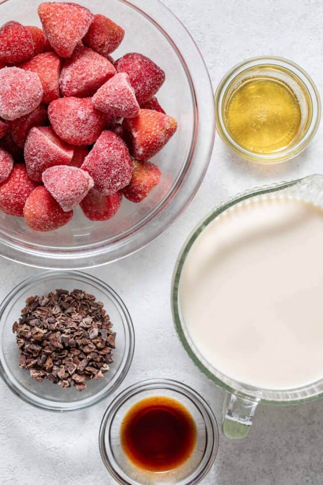 Clear mixing bowls filled with frozen strawberries, cacao nibs, pure vanilla extract, oat milk and honey.