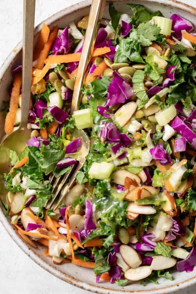 A tan colored bowl filled with chopped kale salad (curly kale with cabbage, carrots, celery, nuts, seeds and raisins).
