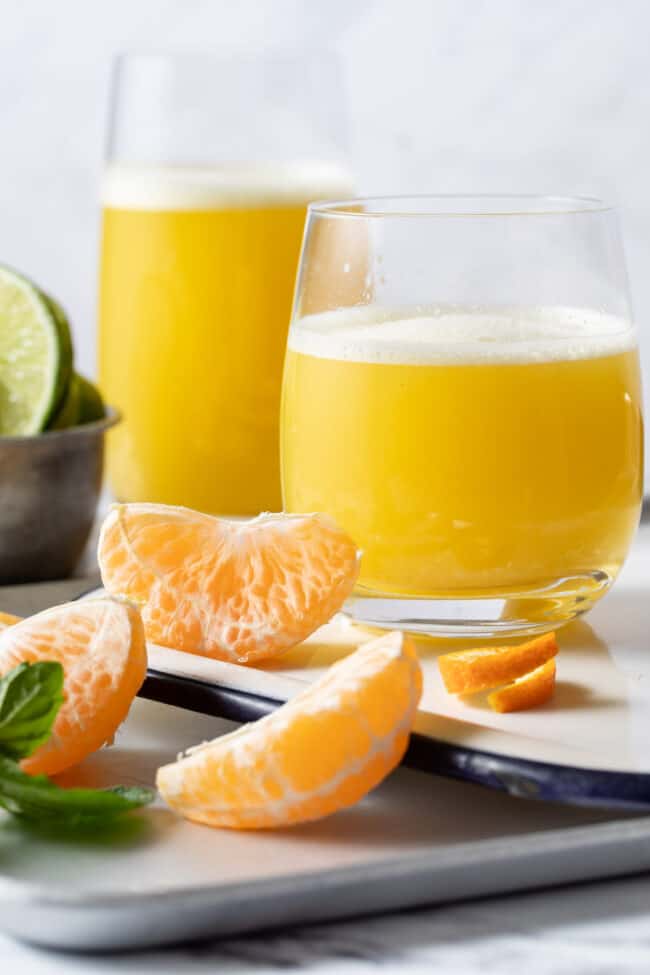 Two glasses on a white tray are filled with Orange Ginger Turmeric Juice. Peeled orange sections are scattered on the tray next to the glasses.