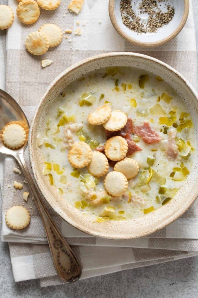 A tan bowl filled with New England Clam Chowder. The soup is garnished with pieces of cooked bacon and Oyster crackers. A spoon sits next to the bowl.