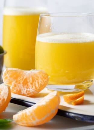 Two glasses filled with Orange Ginger Juice sits on a white tray. Peeled orange sections sit next to the glasses.
