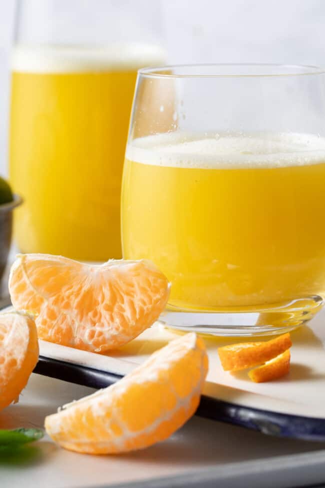 Two glasses filled with Orange Ginger Juice sits on a white tray. Peeled orange sections sit next to the glasses.