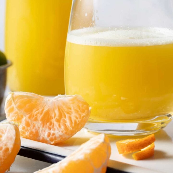 Two glasses filled with Orange Ginger Juice sits on a white tray. Peeled orange sections sit next to the glasses.